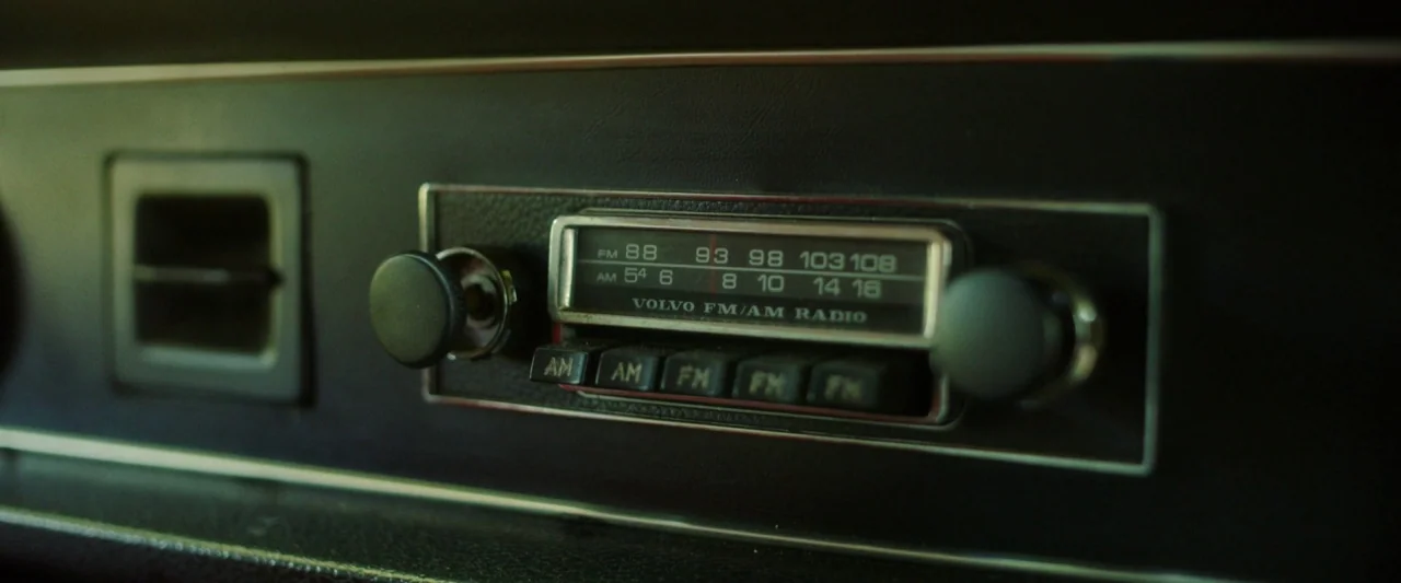 Stockholm (2019) movie still: closeup — A close-up of a vintage Volvo FM/AM radio mounted in a textured black dashboard. The radio dial and…