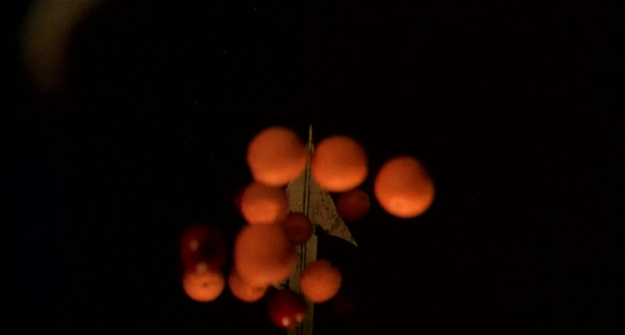 Raising Cain movie still: closeup — A group of glowing, orange-hued spheres floats against an absolute black void. A sharp metallic sliver sits…