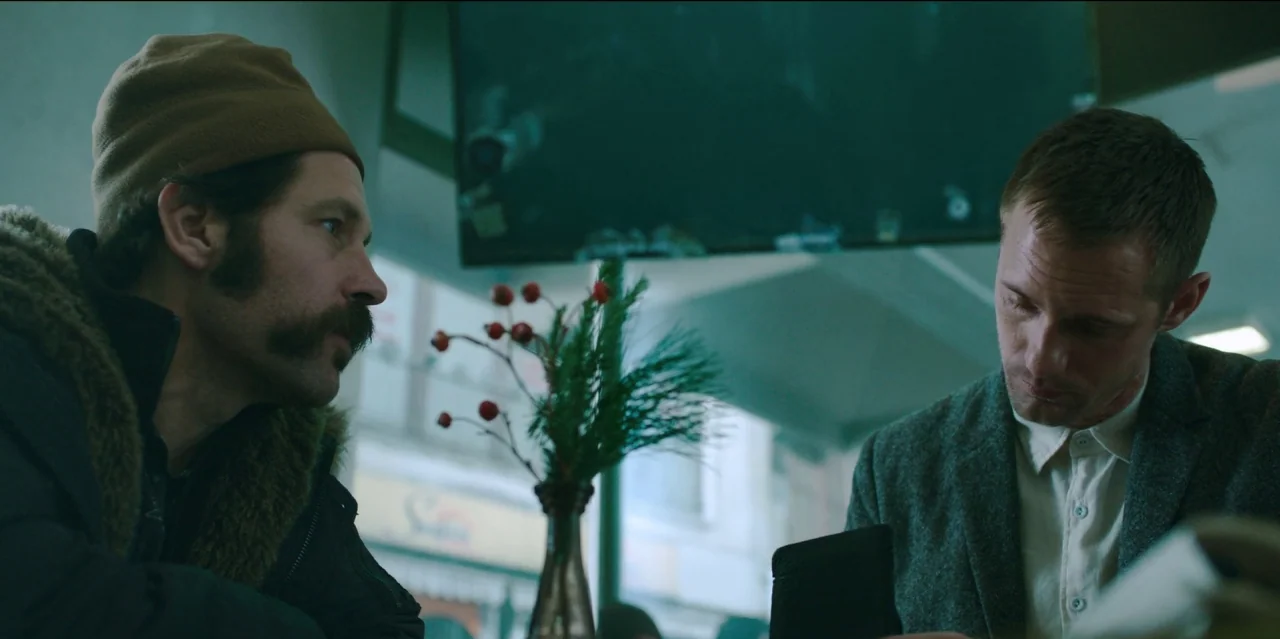 Mute (2018) movie still: medium — Two men sit across from each other at a booth near a bright window. A small vase with red berries sits between…