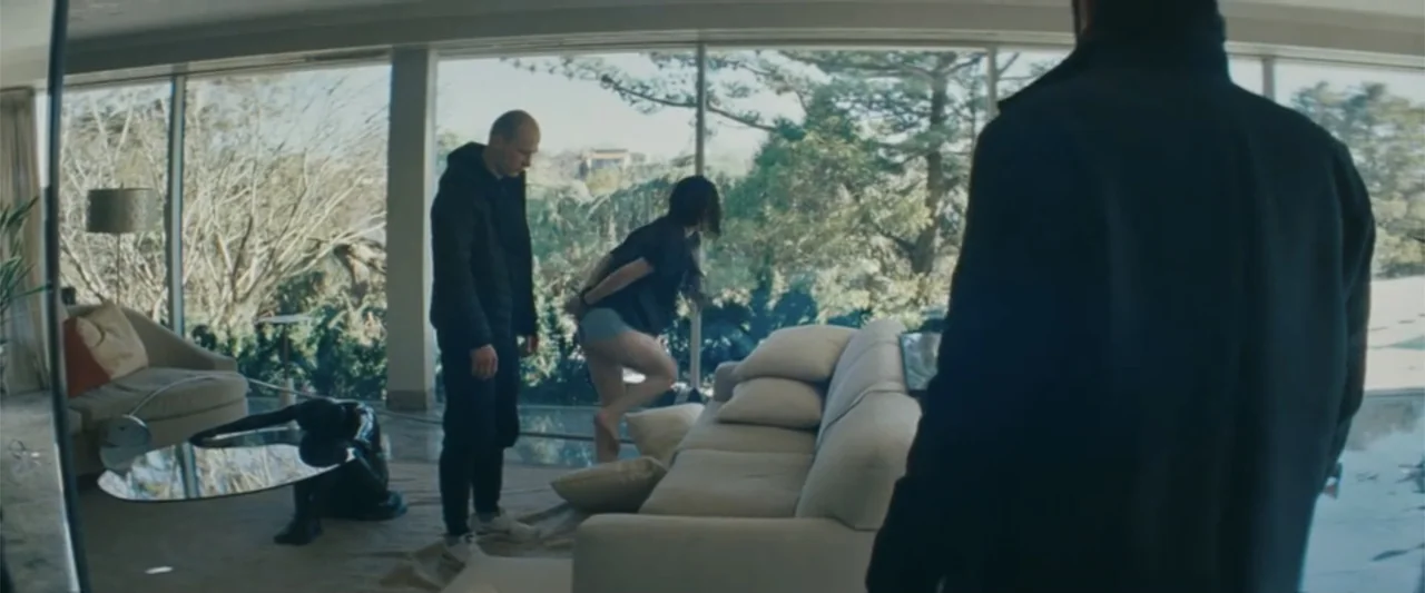Anora (2024) movie still: fullbody — A minimalist room features floor-to-ceiling windows overlooking trees and a large sofa. A bald man stands…