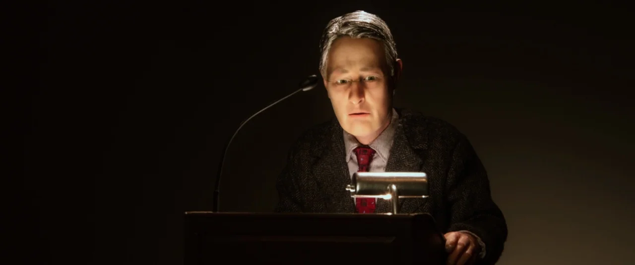 Anomalisa (2015) movie still: medium — A man stands behind a dark wooden podium in a pitch-black environment. He looks down at a document while a…