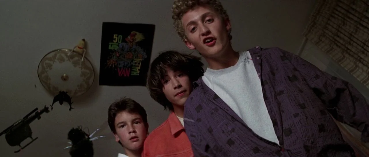 Bill and Ted’s Excellent Adventure (1989) movie still: medium — A dimly lit room decorated with posters and hanging toy helicopters. Three teenage…