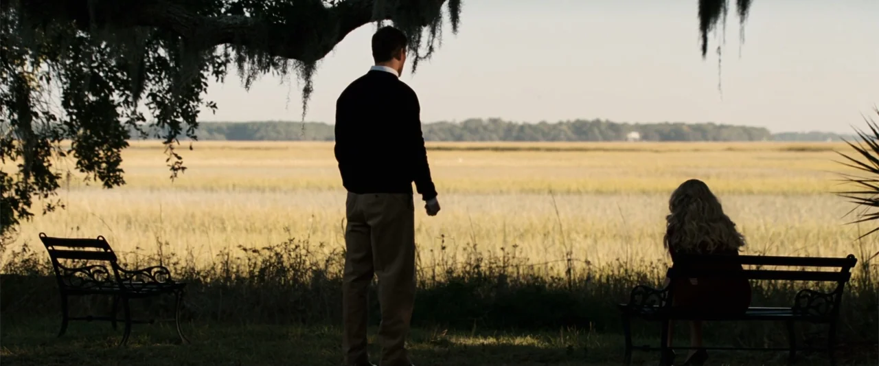 Dear John (2010) movie still: fullbody — An open marshland landscape features two black metal benches under a large oak tree. A man standing and a…