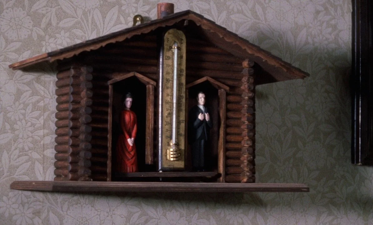 Dr Jekyll & Sister Hyde (1971) movie still: closeup — A wooden weather house barometer is mounted on a wall with floral wallpaper. Two small plastic…
