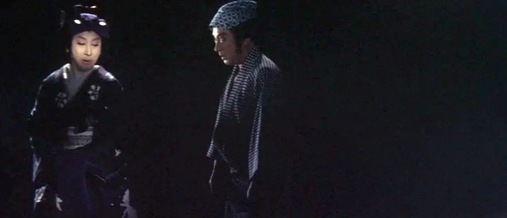 An Actor’s Revenge (1963) movie still: medium — Two people stand in the center of a pitch-black, empty theatrical stage space. They are illuminated…