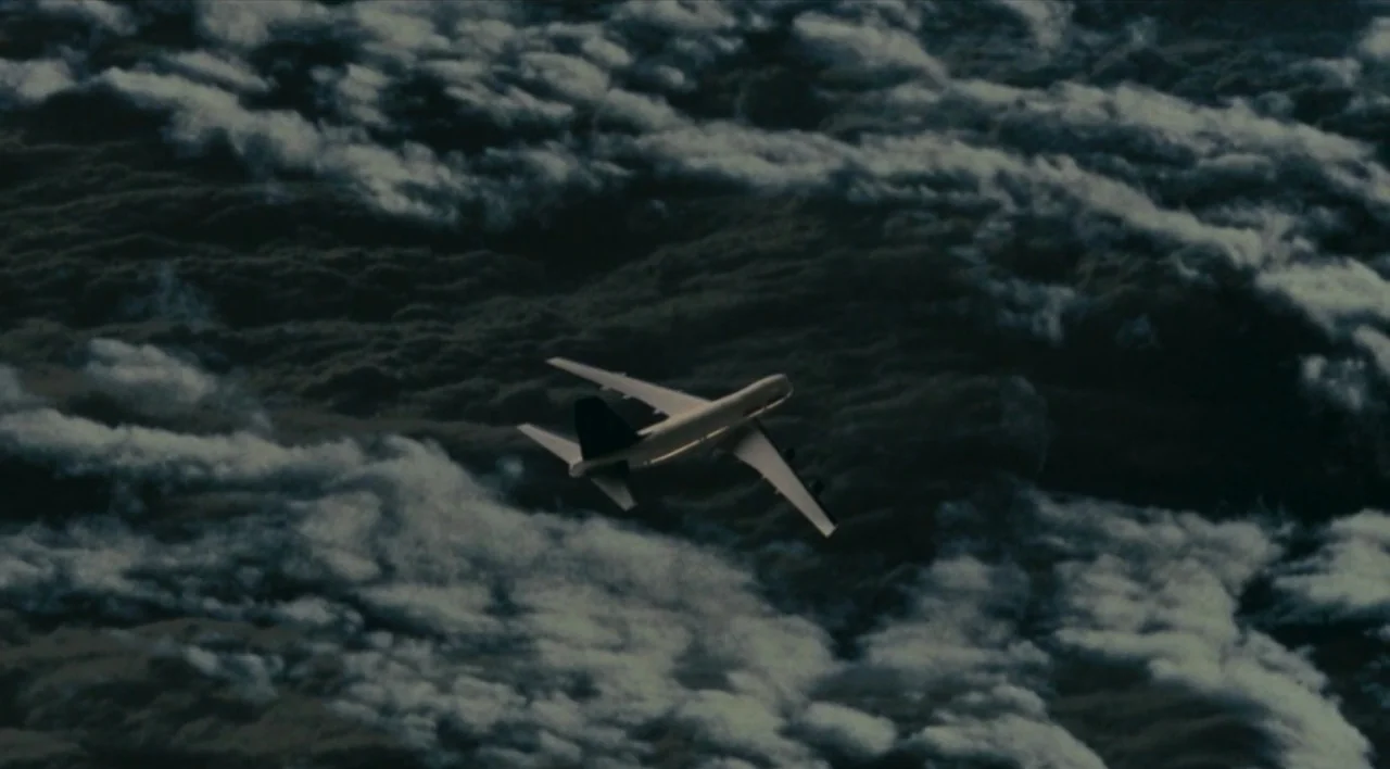The Other Conquest (1999) movie still: wide — A wide view of a commercial jet flying high above a dense layer of dark clouds. The aircraft moves…