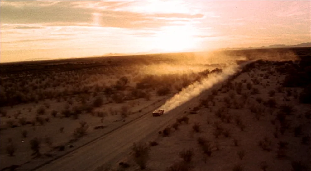 U-Turn (1997) movie still: establishing — A vast, flat desert landscape stretches toward a horizon beneath a golden sunset. A single vehicle travels…