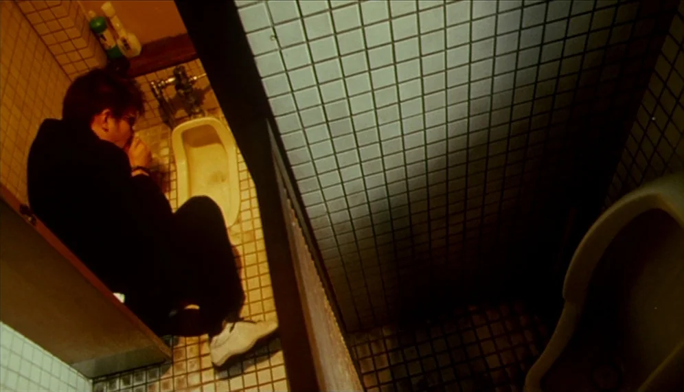 Gozu (2003) movie still: medium — A small, tiled restroom stall features a squat toilet and white ceramic grid tiling. A man sits on the floor with…