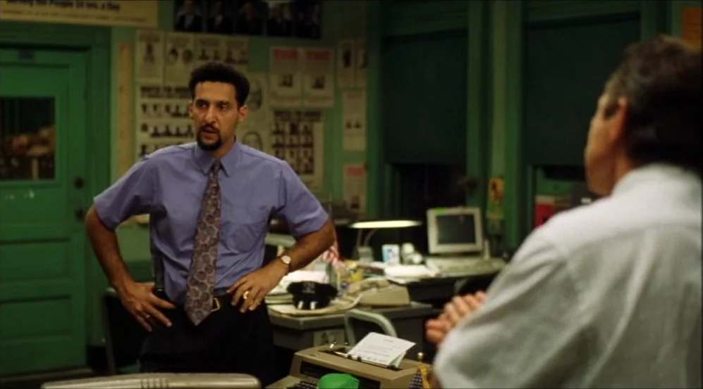 Clockers (1995) movie still: medium — A green-walled office features bulletin boards covered in headshots and papers. A man in a blue shirt stands…