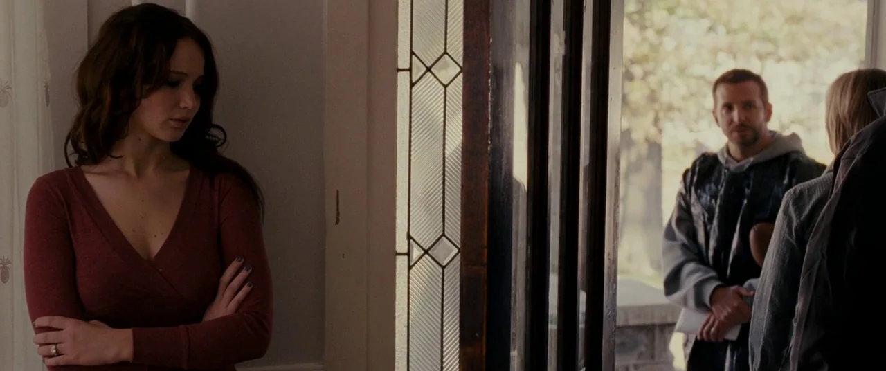Silver Linings Playbook (2012) movie still: medium — A woman stands inside by a glass-paned door looking toward an outdoor entry. A man stands on…