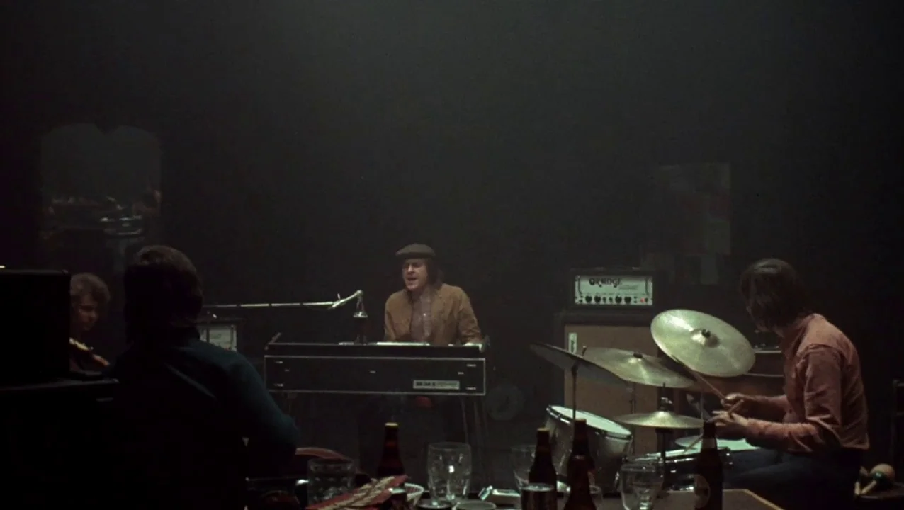 O Lucky Man! (1973) movie still: wide — A dimly lit recording studio is filled with a soft haze and dark shadows. A band is practicing music while…