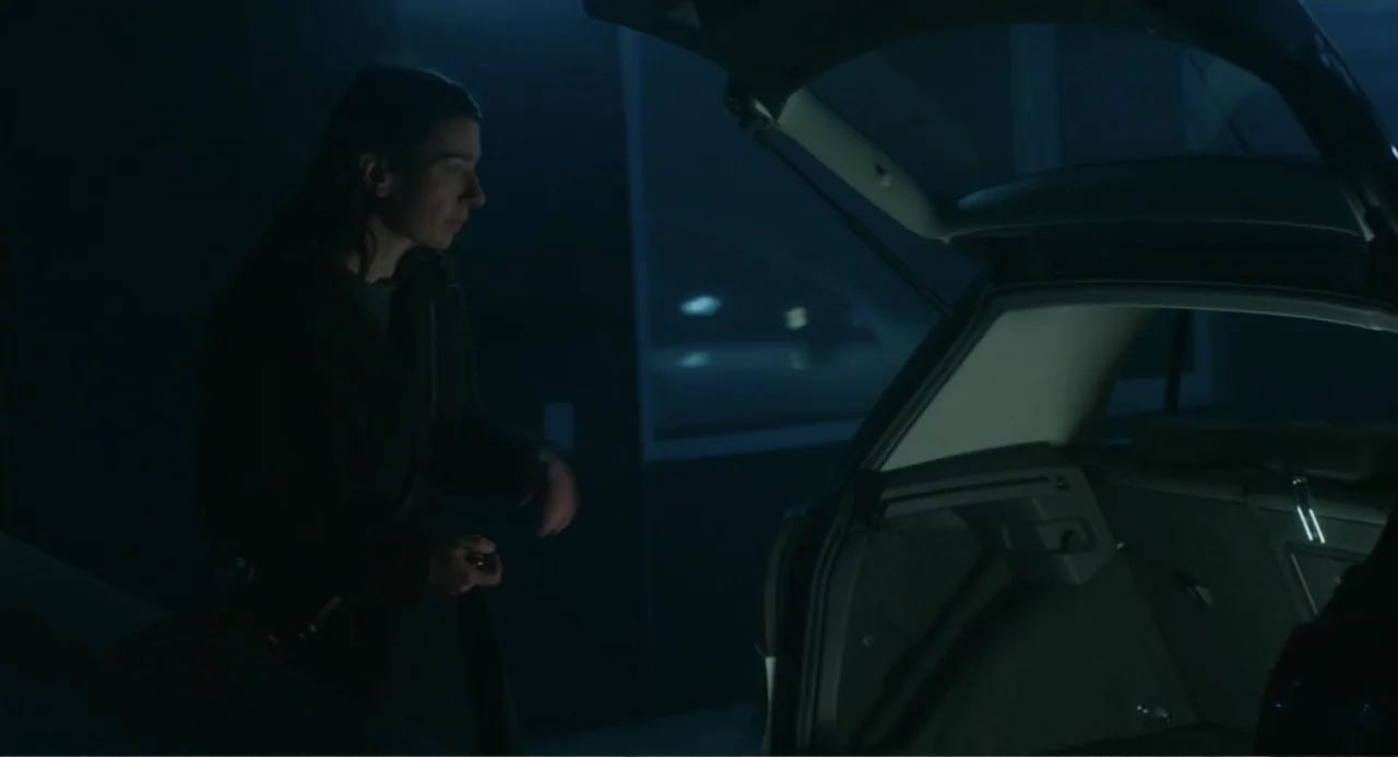 Orphan: First Kill (2022) movie still: medium — A dimly lit parking garage at night contains a parked car with an open trunk. A woman in a dark coat…