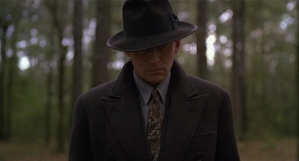 Miller’s Crossing (1990) movie still: medium — A man stands in a dense, green forest with vertical trees blurred in the distance. The man wearing a…