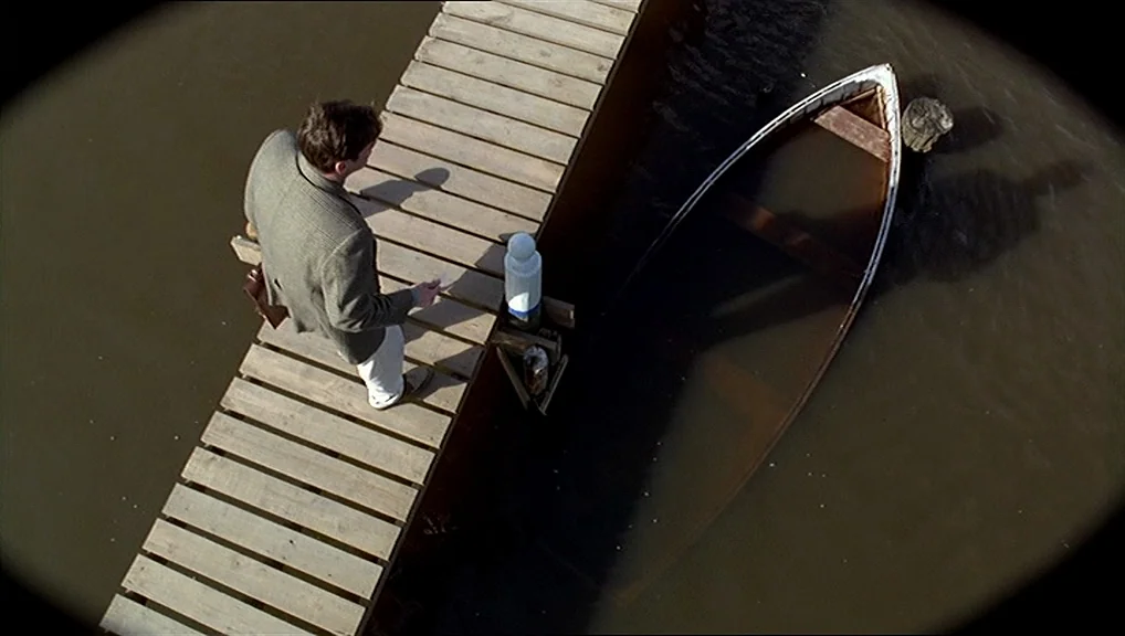The Truman Show movie still: wide — A wooden plank pier extends into murky brown water next to a half-sunken boat. A man in a light suit stands on…