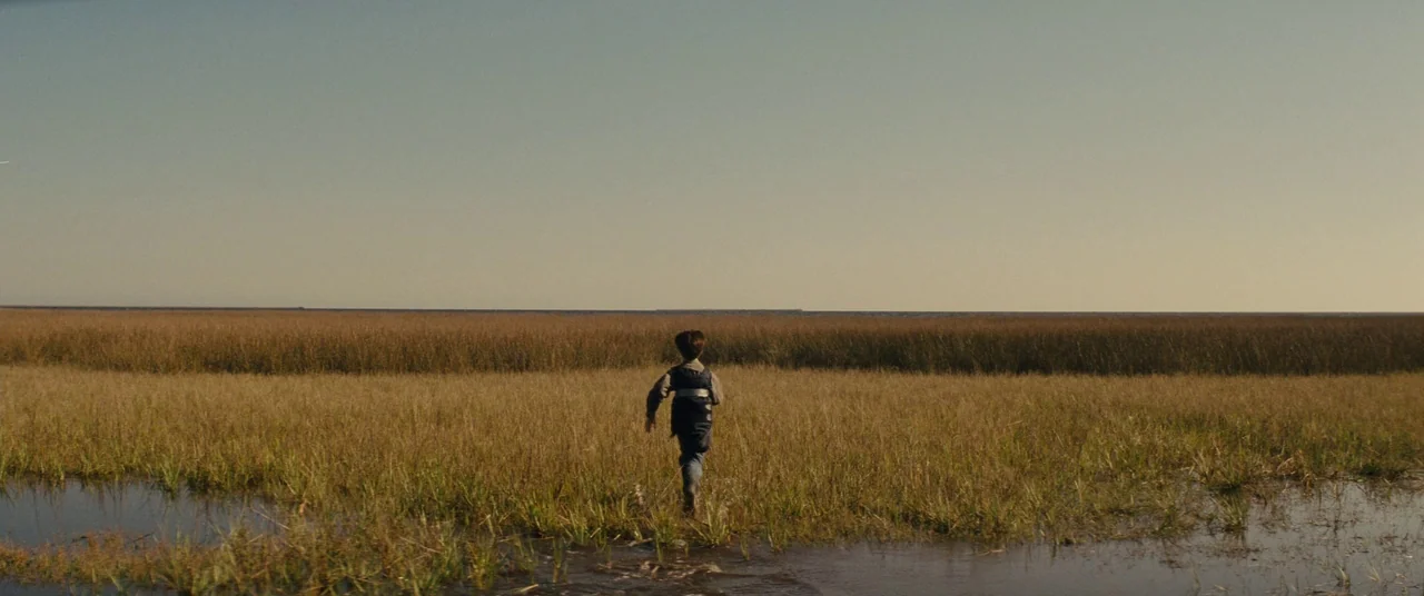 Midnight Special (2016) movie still: wide — A vast, flat marshland stretches to a distant horizon line under a pale sky. A lone young boy runs…