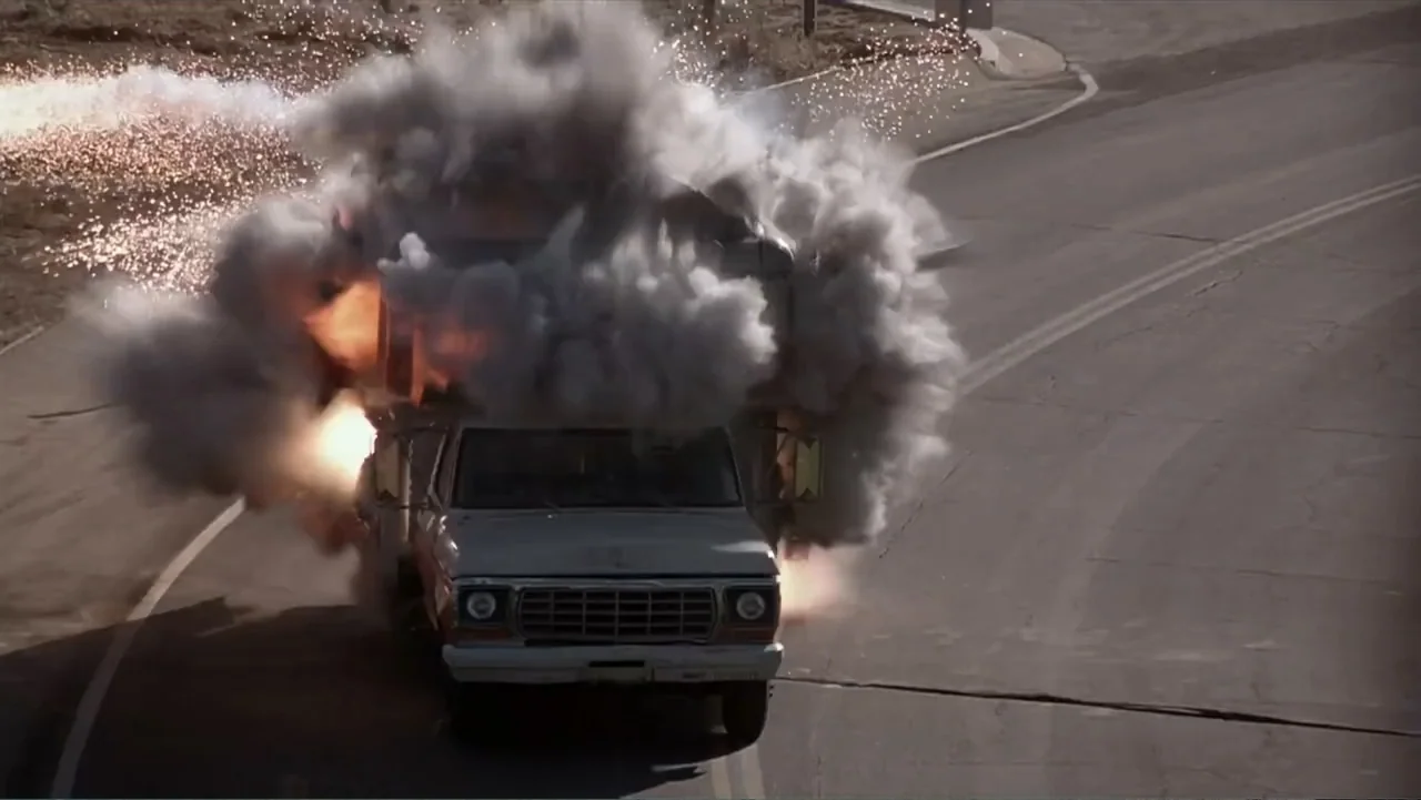 Crisis Point (2012) movie still: wide — A white recreational vehicle travels down a winding asphalt road during the day. A large explosion of grey…