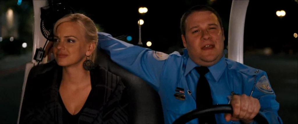 Observe And Report (2009) movie still: medium — A man in a blue security uniform drives a small vehicle at night. A woman with blonde hair and round…