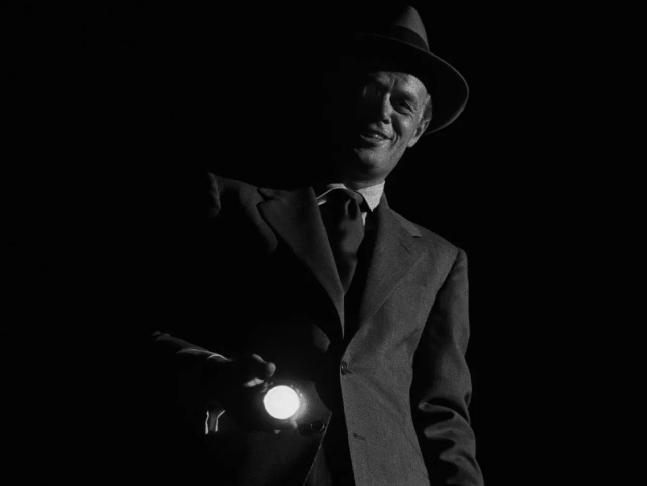 Pickup on South Street (1953) movie still: medium — A man stands in an indeterminate dark space wearing a 1940s style suit. He points a bright…