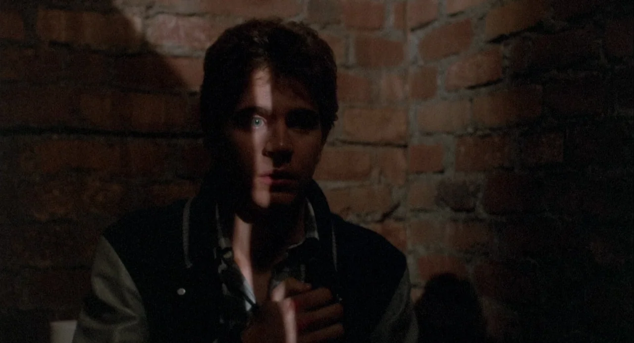 Vamp (1986) movie still: medium — A dark corner with a raw, exposed brick wall serves as the backdrop. A young man sits in the shadows with part of…