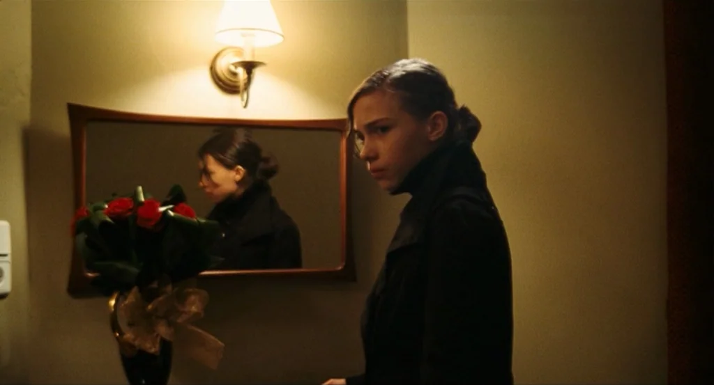 This Night (2008) movie still: medium — A dim hallway features a wall-mounted mirror above a bouquet of red roses. A young woman in a dark coat…