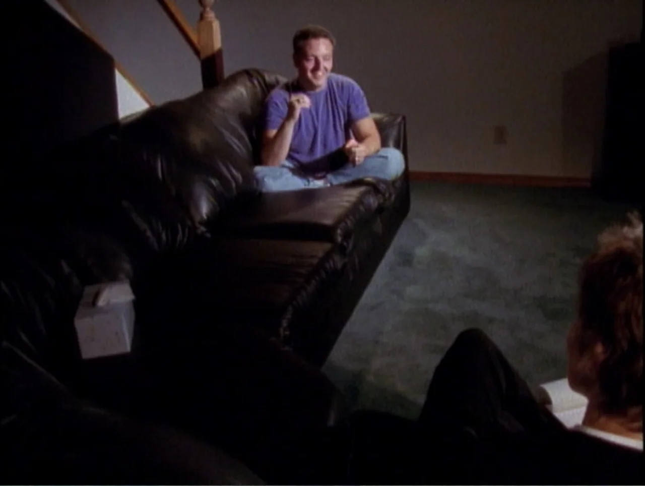 America Undercover: Life Afterlife (2000) movie still: medium — A basement living room features a dark leather couch and teal carpeting. A man sits…