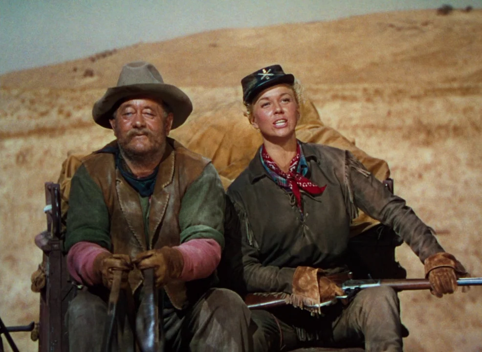 Calamity Jane (1953) movie still: medium — An older man and a blonde woman sit side-by-side in a horse-drawn wagon. The background shows a dry,…