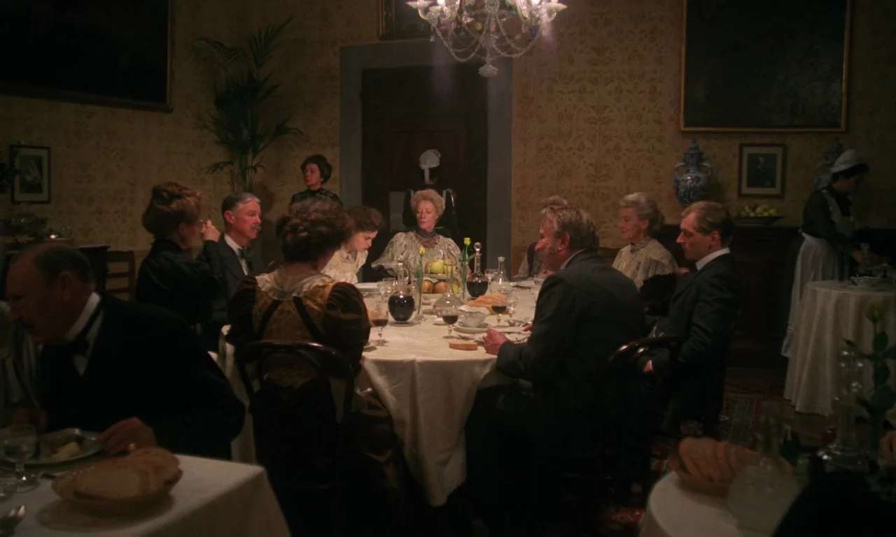A Room With A View (1985) movie still: wide — An opulent, dimly lit room with ornate wallpaper hosts an formal dinner party. Multiple guests in…