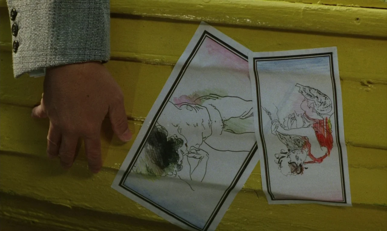 Yumeji (1991) movie still: closeup — Two ink and colored pencil sketches lie on bright yellow horizontal wooden planks. A hand wearing a grey jacket…