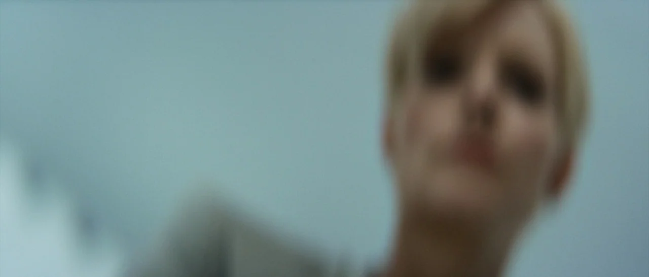Elysium (2013) movie still: closeup — A featureless indoor space is presented with a soft, out-of-focus background. A blonde woman looks directly…