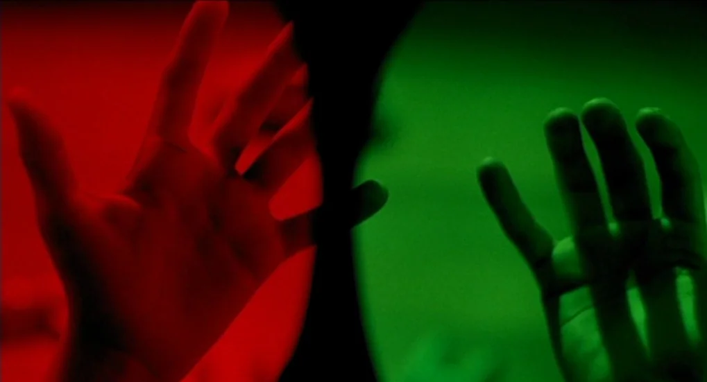 Jeux d’Enfants (2003) movie still: closeup — An abstract dark space is split vertically into red and green halves. Two disembodied hands reach…