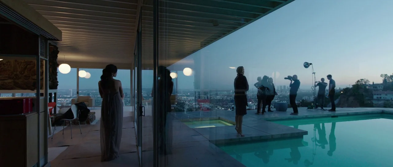 Knight of Cups (2015) movie still: wide — A modern glass-walled architectural home overlooks a sprawling urban landscape at twilight. A woman stands…