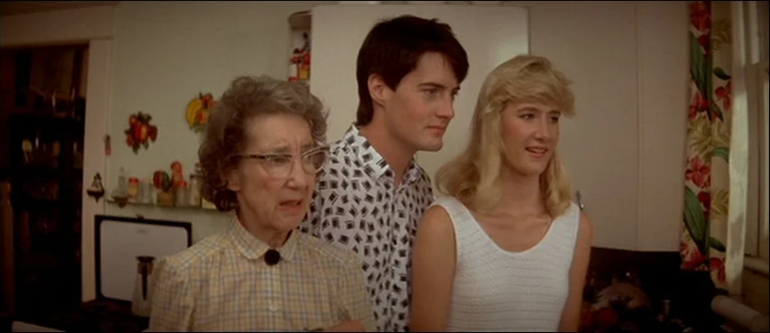 Blue Velvet (1986) movie still: medium — Three people stand together in a kitchen with retro wallpaper and decorative wall hangings. They look…