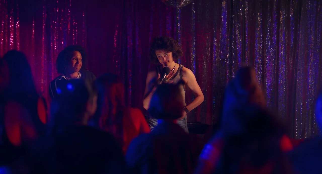Drive-Away Dykes (2024) movie still: medium — A dark performance venue features shimmering sequined curtains reflecting pink and blue light. One…