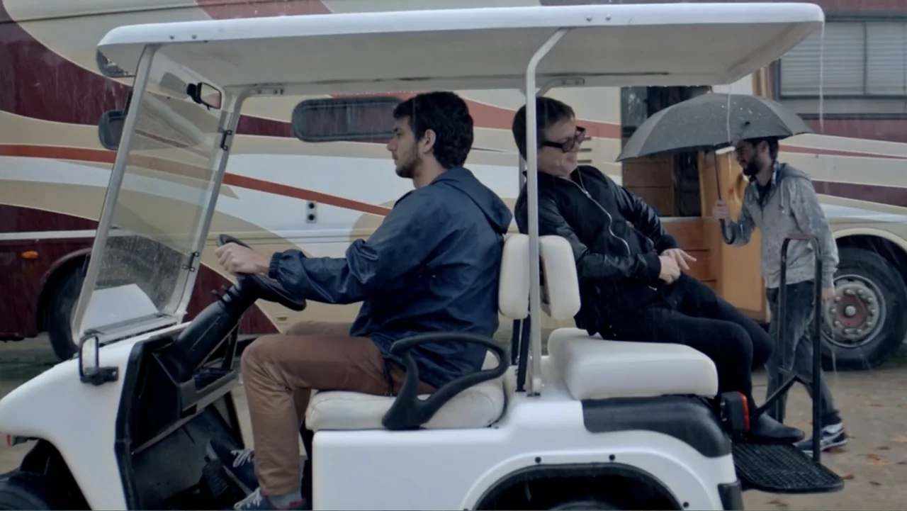 I Married a Dumbass (2016) movie still: medium — A rain-dampened parking lot with a large recreational vehicle parked in the background. Two men sit…