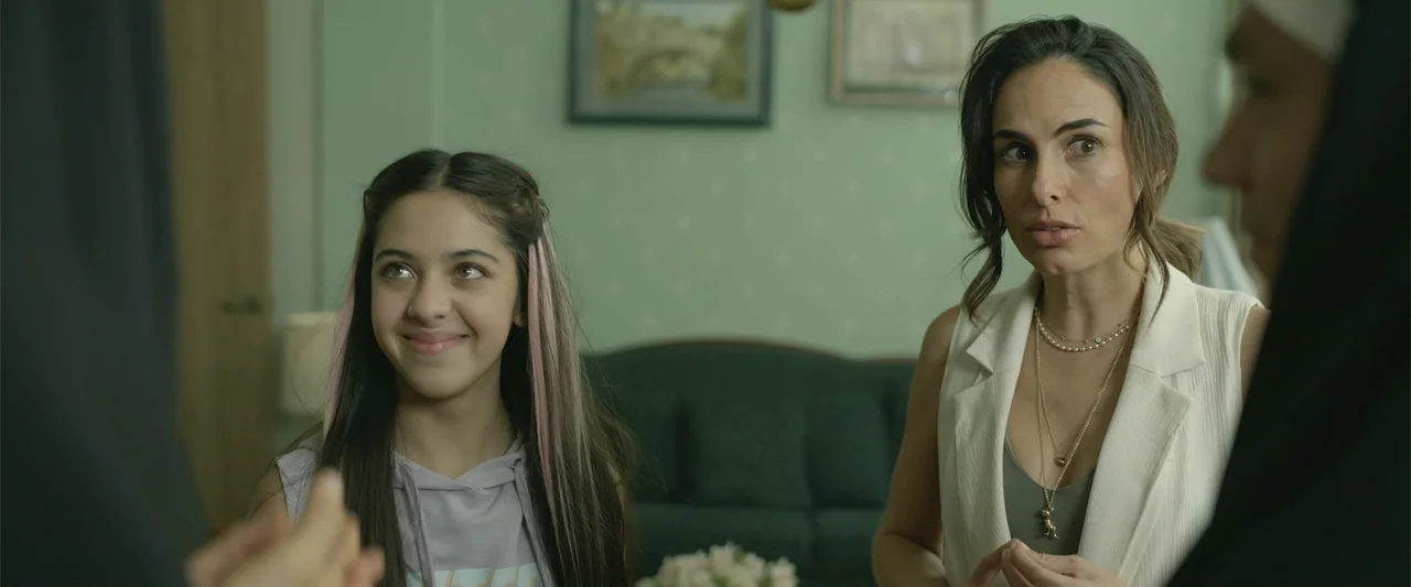 Divine Intervention (2026) movie still: medium — A living room has pale green walls decorated with framed pictures and a sofa. A girl smiles at a…