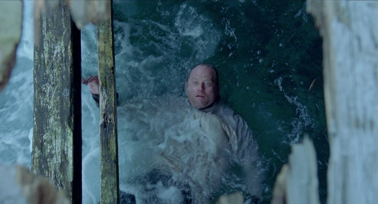 Insomnia (1997) movie still: medium — A man treads water beneath the remnants of a broken wooden pier. Rushing water surrounds the man while he…