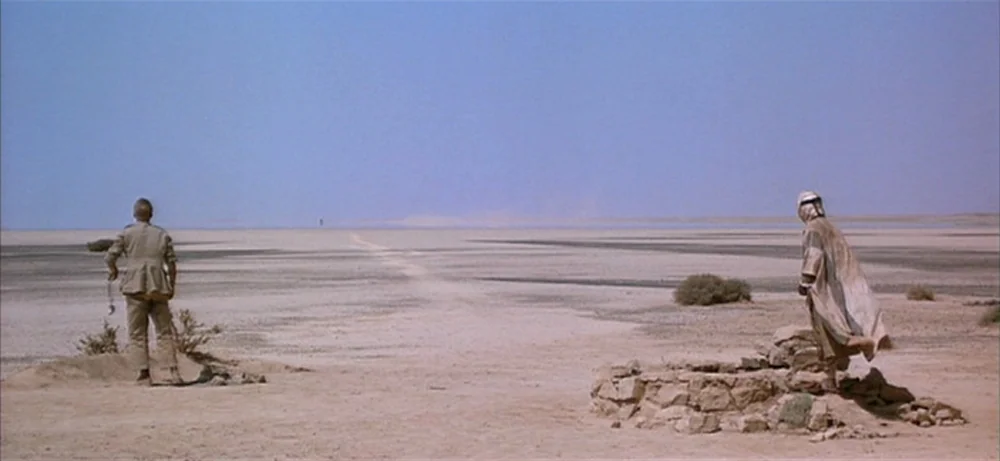 Lawrence Of Arabia movie still: wide — A vast, flat desert landscape stretches under a clear sky. Two men stand apart near a small circular stone…
