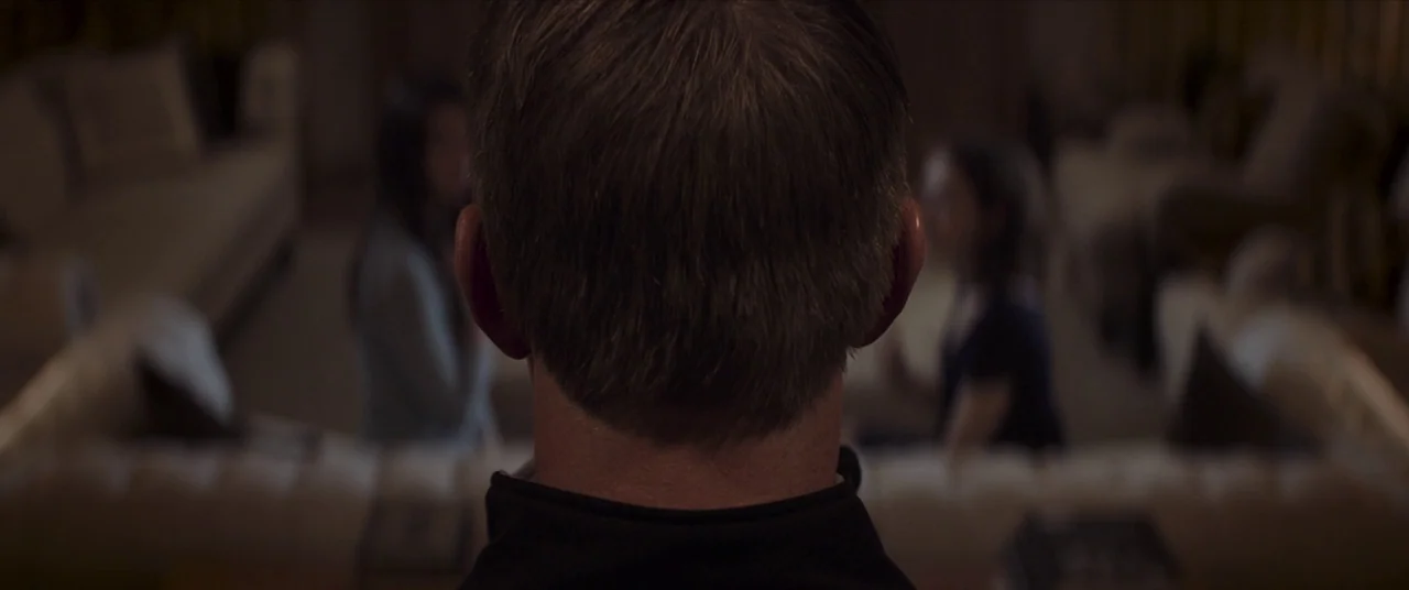 Thoroughbreds (2017) movie still: closeup — A dimly lit room with a person in the immediate foreground facing away. Two other figures are blurred in…