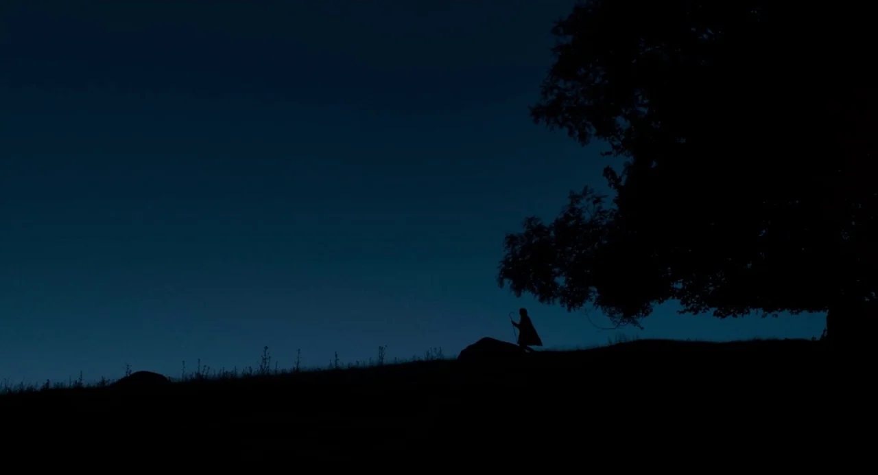Dark River (2017) movie still: wide — A dark, grassy hill rises against a deep blue evening sky. A single hooded figure holding a staff walks across…