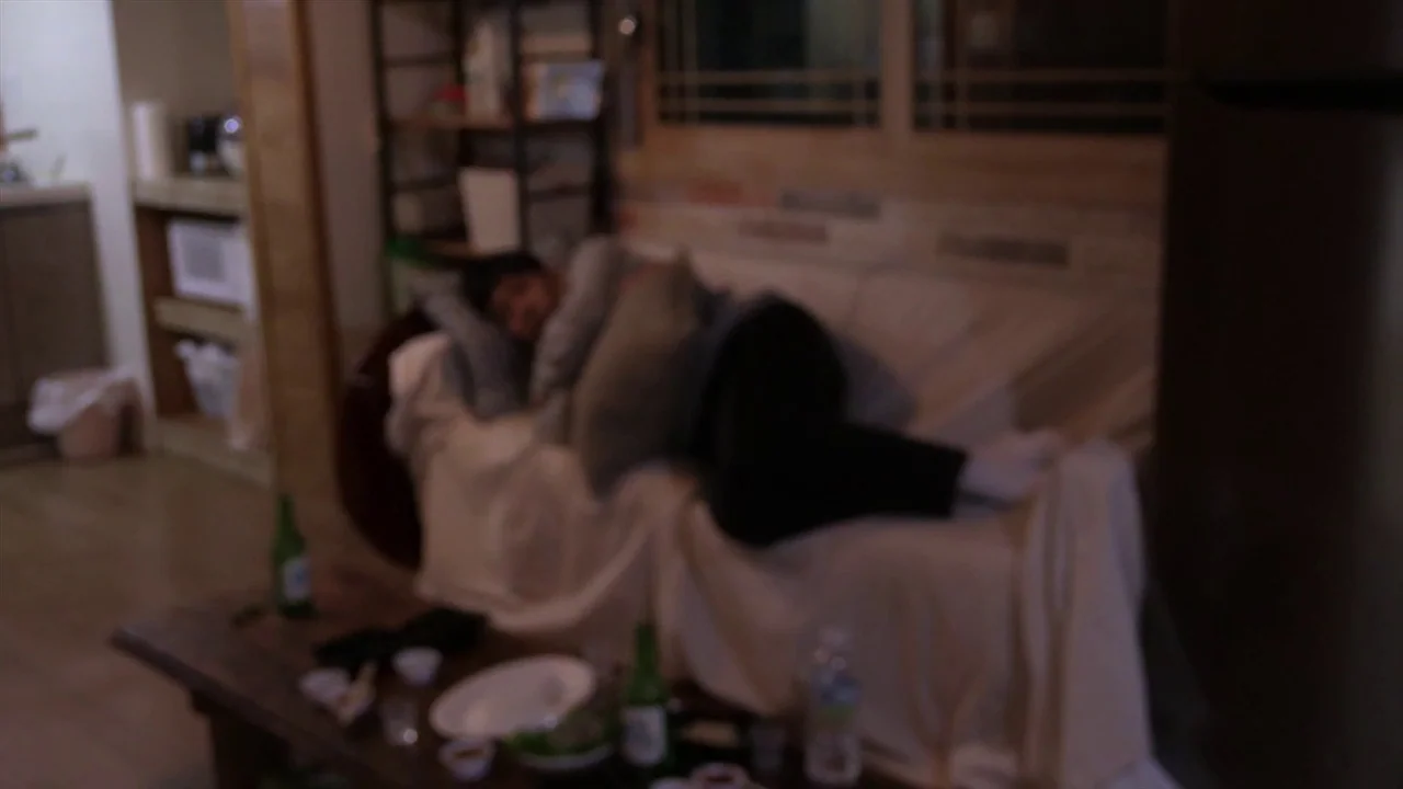 In Water (2023) movie still: medium — A dimly lit room features a couch draped in a light-colored cloth cover. A person sleeps in a fetal position…