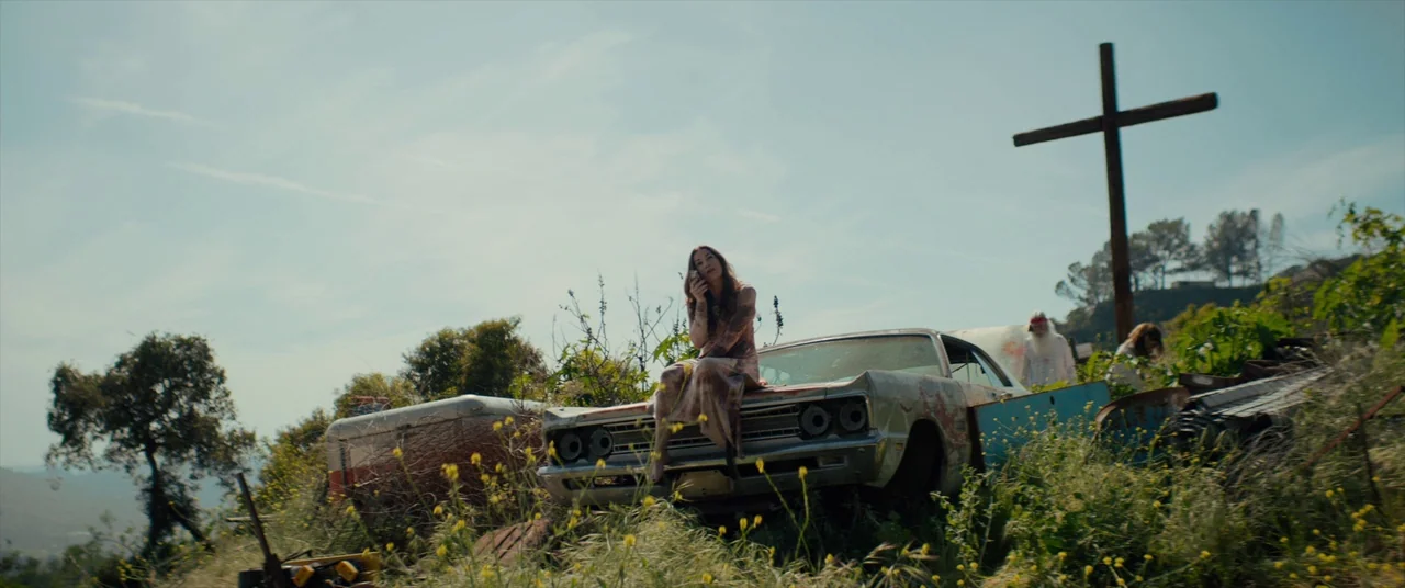 Tone Deaf (2019) movie still: wide — A desolate hillside filled with overgrown weeds and abandoned, rusted vehicle parts. A woman sits on the hood…