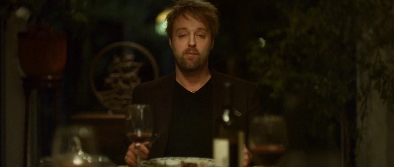 Depraved (2019) movie still: medium — A dimly lit dining room contains a person seated at a table. The man sits alone with a meal and wine in front…