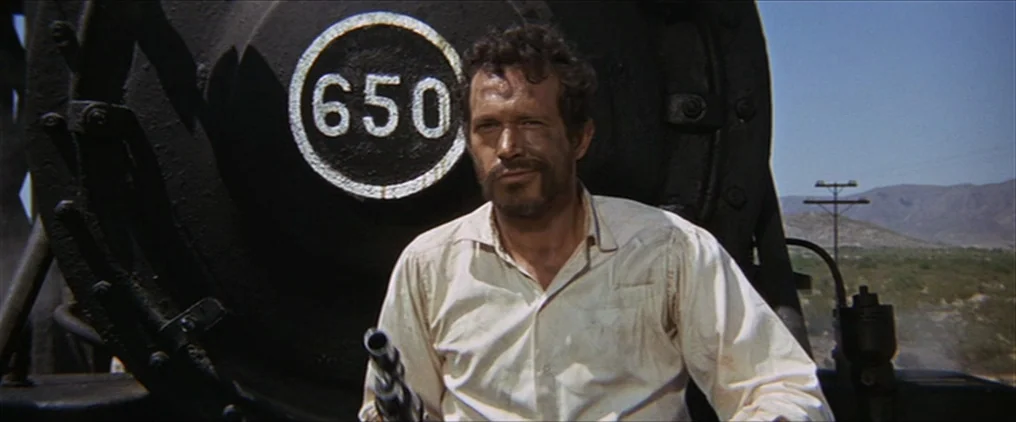 The Wild Bunch (1969) movie still: medium — A man sits in front of a large black iron train locomotive numbered 650. He holds a pistol while looking…