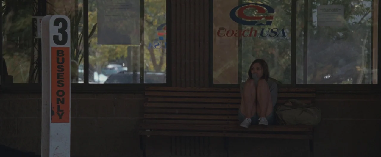 Martha Marcy May Marlene (2011) movie still: wide — A bus station exterior features a wooden bench beneath glass windows with Coach USA signage. A…