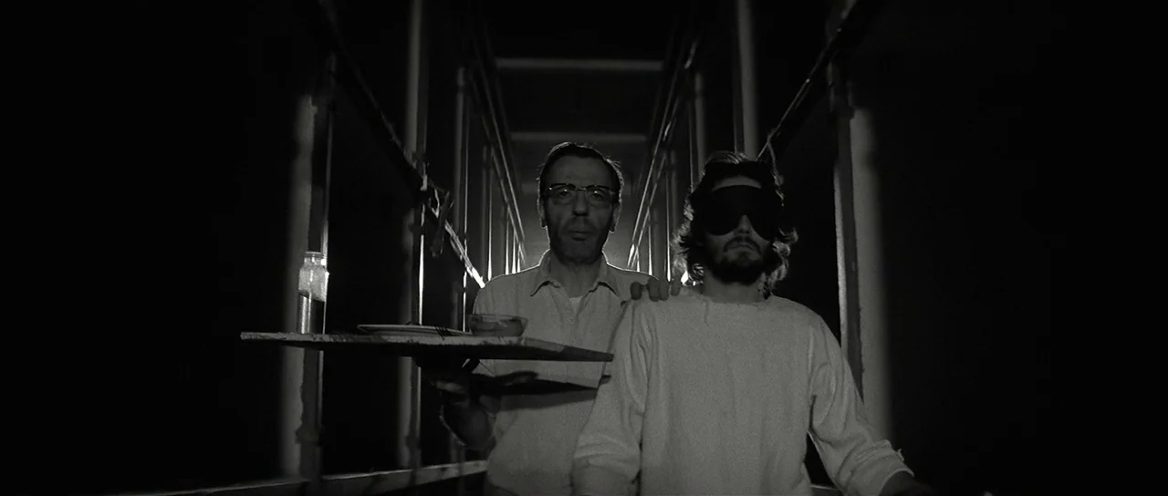 The Last Battle (1983) movie still: medium — An enclosed, shadowed narrow walkway with vertical metal supports disappears into darkness. A man…