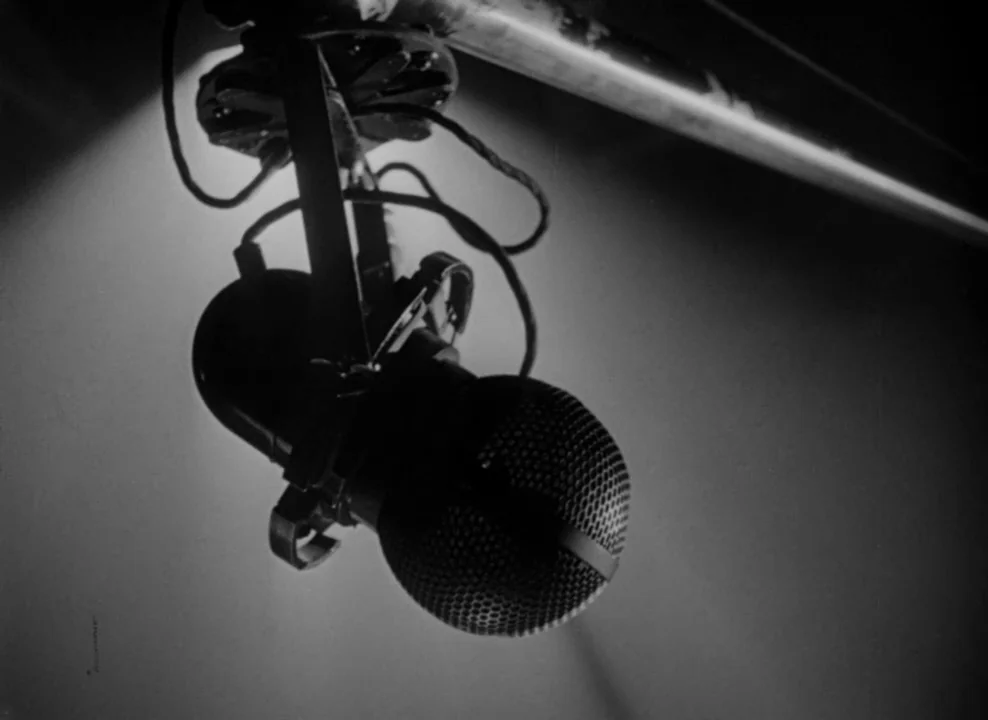 The Magnificent Ambersons (1942) movie still: closeup — A vintage metal microphone hangs suspended from an overhead boom arm in darkness. The…