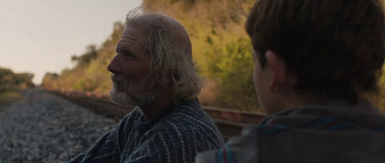 Joe (2013) movie still: medium — An elderly man and a boy sit beside overgrown railroad tracks at sunset. The pair occupies the foreground while a…