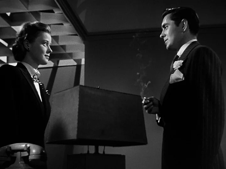 Nightmare Alley (1947) movie still: medium — A man and a woman stand on opposite sides of a desk in a dark room. The man holds a cigarette as thin…