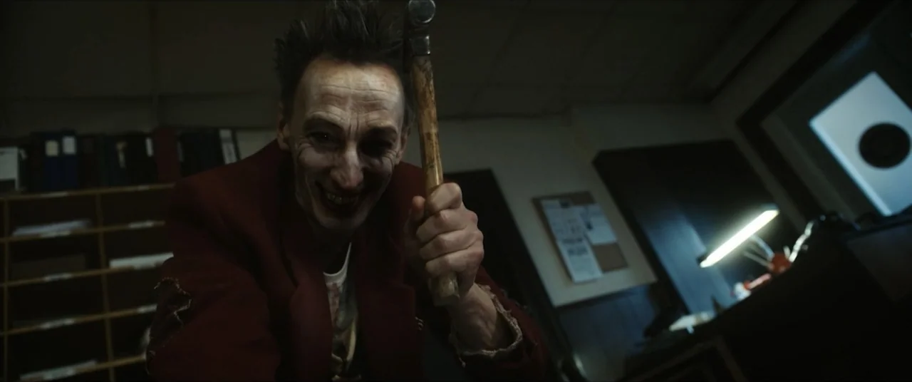 Vicious Fun (2021) movie still: medium — An office at night contains dark wooden shelving and a lamp on a desk. A thin man in a red blazer looms…