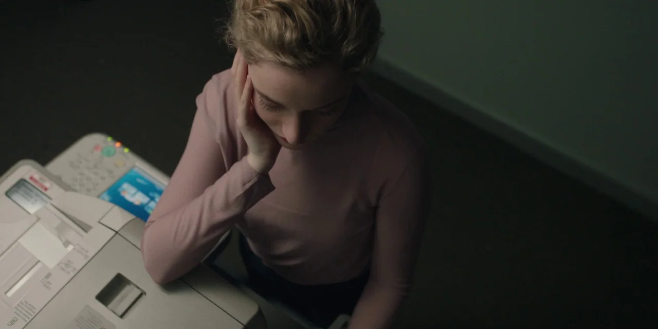 The Assistant (2019) movie still: medium — A dimly lit office features a woman standing next to a large photocopier. She rests her head on her hand…